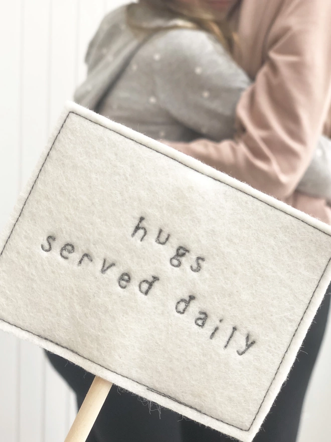 hugs served daily embroidered felt sign with people hugging in background
