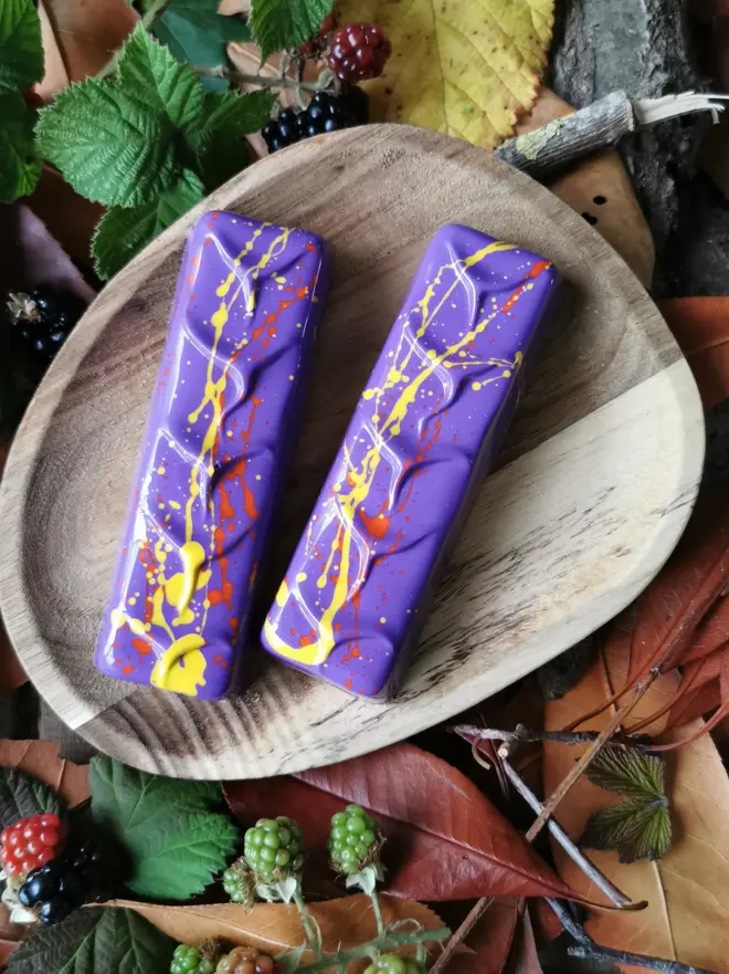 Two purple choclate bars with red and yellow splatter designs, sitting on a wooden plate resting on fall leaves
