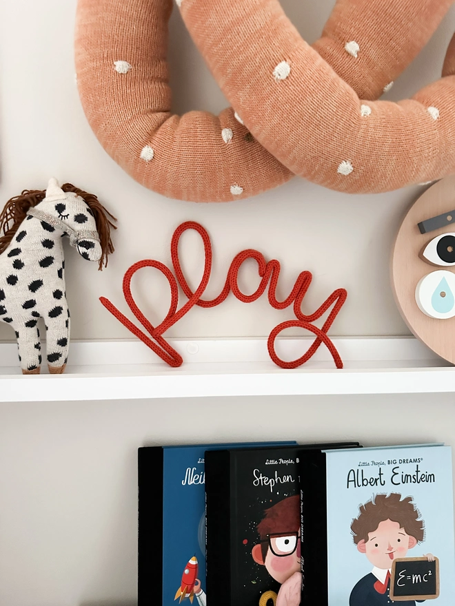 A play knitted word sign on a shelf in a kids playroom with other children's wall decor and kids educational books. 