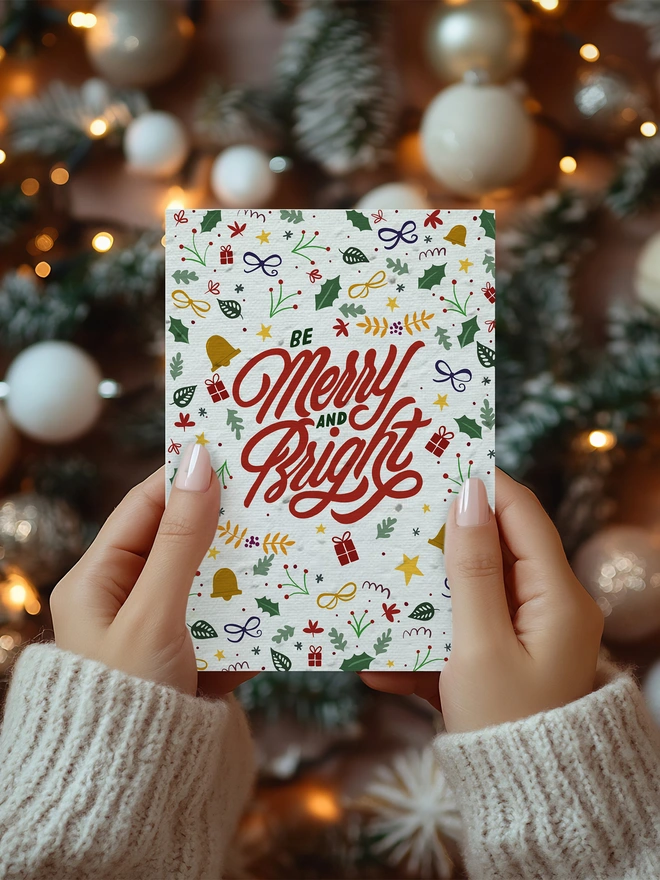 A pair of hands holding a Christmas card with illustrated Christmas motifs on with Merry and Bright in the centre
