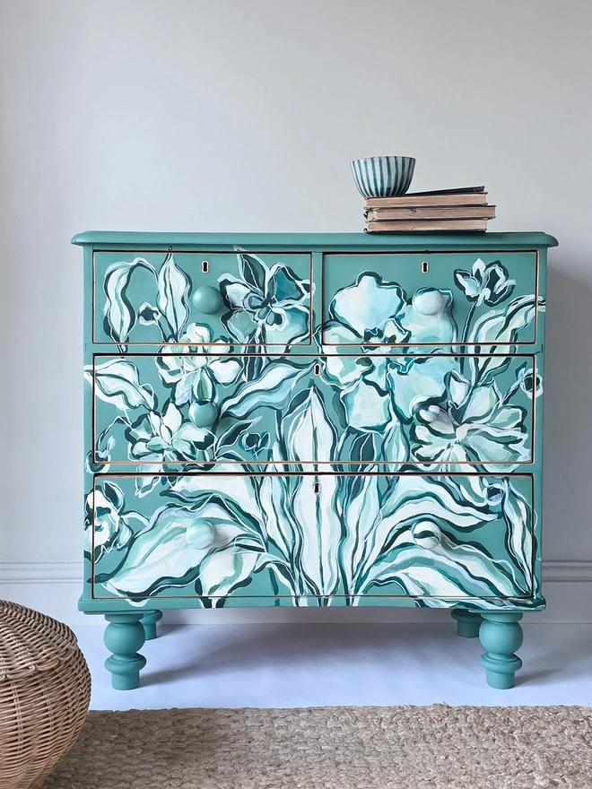 Painted Chest of Drawers staged with a jute rug in the foreground and a lidded basket. On top are vintage books and green striped earthenware; the pine chest of drawers is made from painted rustic pin and has 2 over 3 drawers with victorian wooden knobs. Hand painted with a peony and primrose design in green and cream. 