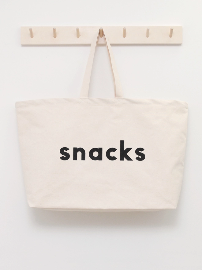 oversized natural canvas tote bag with bold snacks slogan hanging on a wooden hook