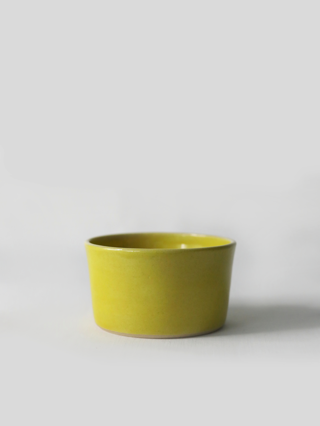 A small handmade ceramic side bowl in glossy sun yellow glaze, with a tapered sides and a wide open rim, photographed against a plain light grey background