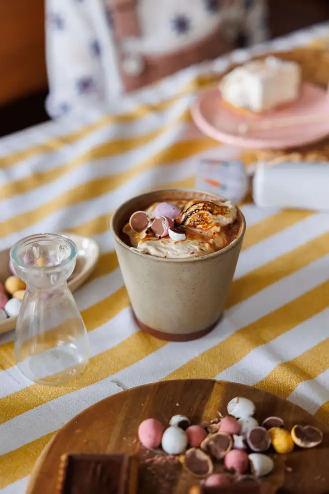 Mini Eggs Wonder Fluff on top of hot chocolate on a striped yellow table cloth surrounded by crushed mini eggs