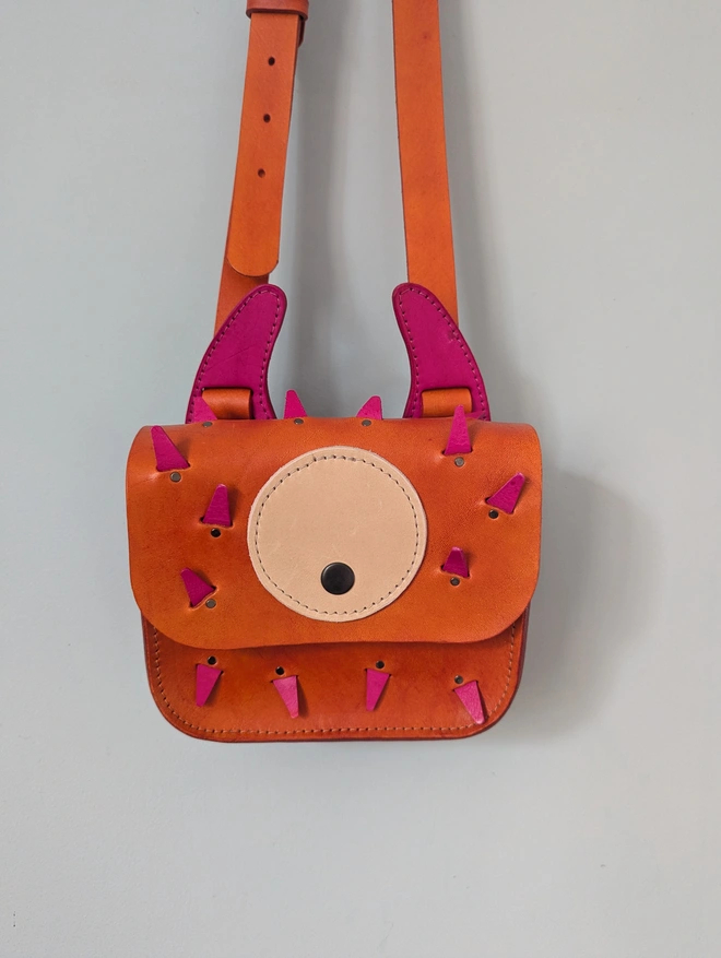 Orange leather monster cross body bag hanging against a blue wall, close view.