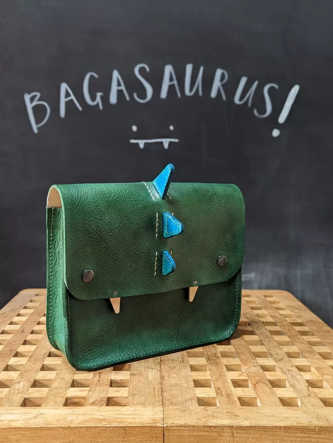 Angled front view of green leather 'Bagasaurus' backpack with turquoise spikes and pointed white fangs