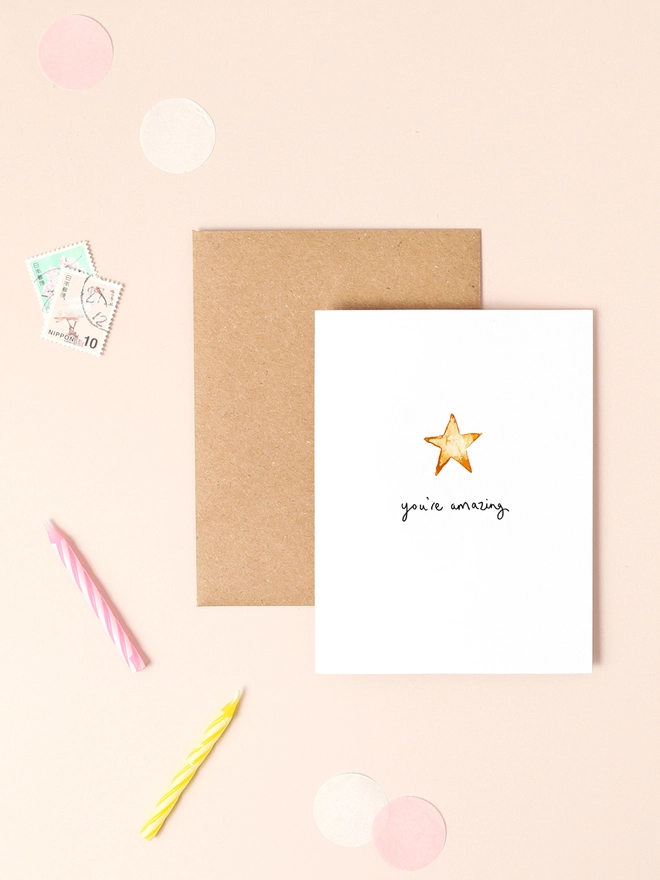 An image of a mini, rectangular white greetings card with an illustration of a gold star with a hand written 'you're amazing' message in black ink seen under the central illustration. The card is seen on top of a brown rectangular kraft envelope.