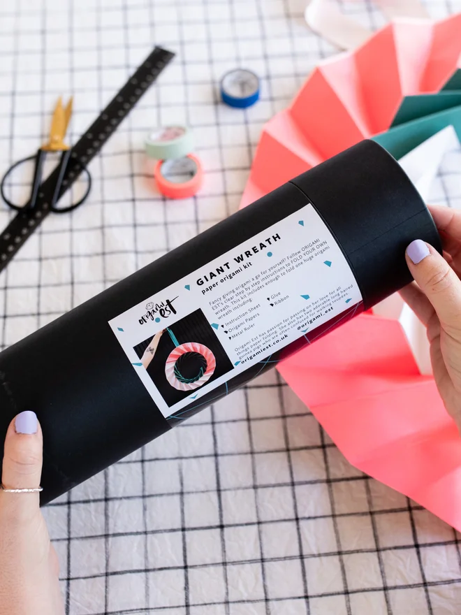 Woman holding an origami kit in a black tube with paper ornament and craft materials in background.