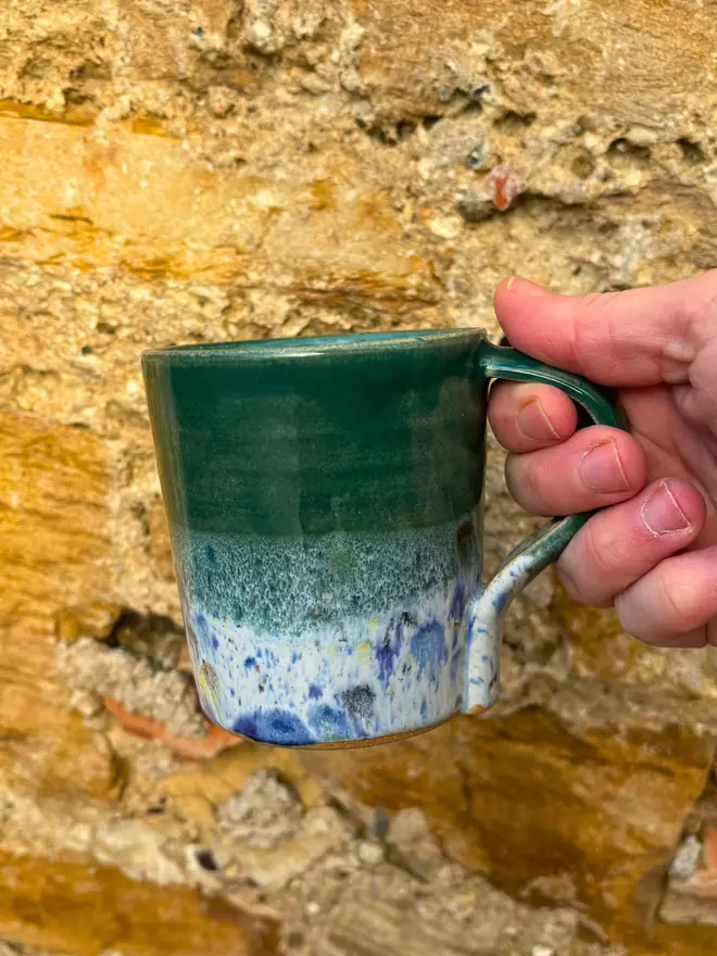 Shoreline Standard Mug, a colourful mug held in front of a brick wall. 