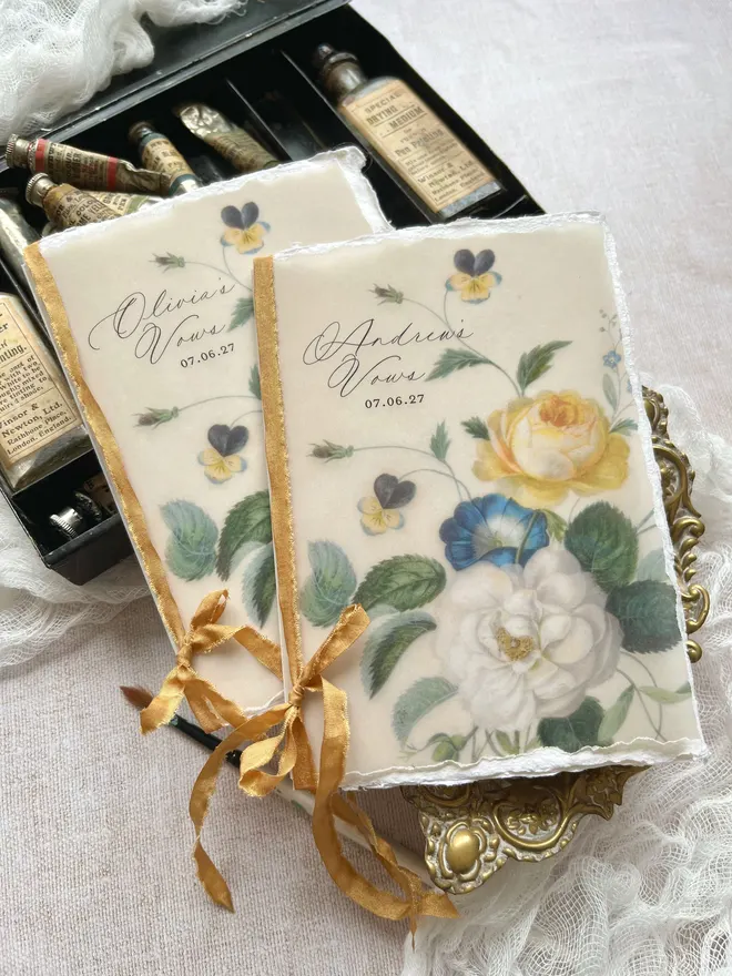 Handmade vow booklets with a silky soft vellum cover printed with yellow, white and blue blooms. The blank pages inside are handmade paper and the pages are all tied together with antique style faux silk ribbon