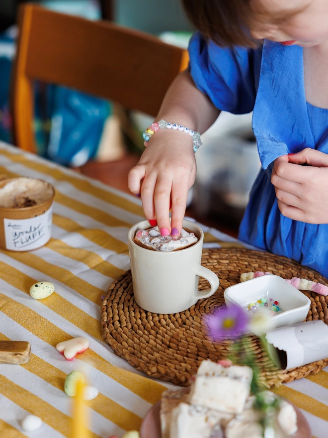child in a blue dress placing a The Mallow Makers jumbo mini egg marshmallow into a mug of hot chocolate on a striped yellow table cloth