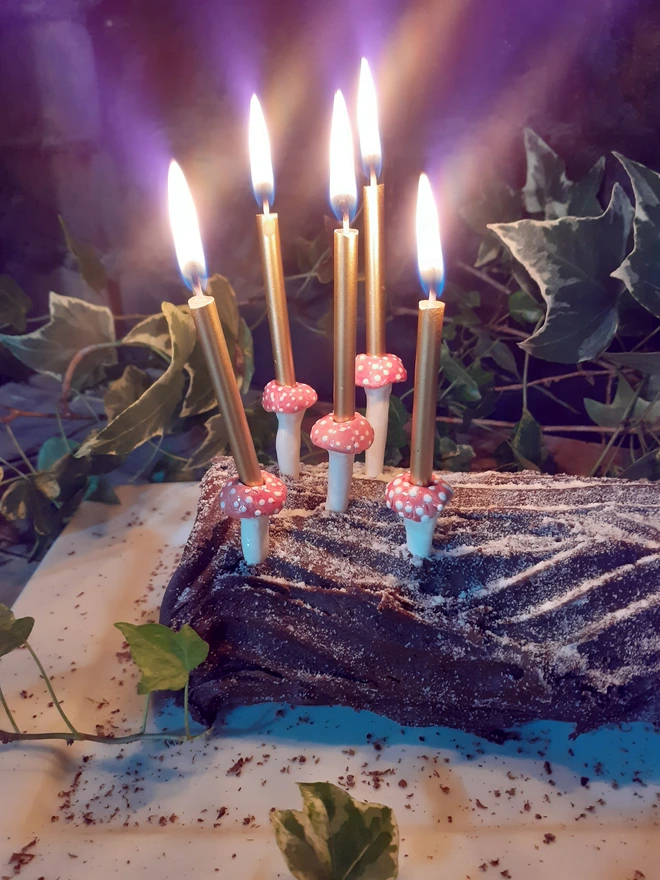 Front view of five ceramic red toadstool candle holders with glowing gold cake candles, arranged across a chocolate log cake in a leafy woodland setting.