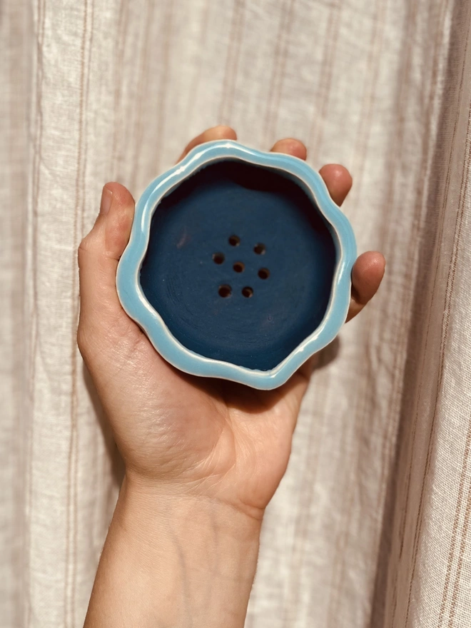 wavy rimmed soap dish held in the palm of a hand against a fabric background. the soap dish is sky blue on the outside and matt dark blue on the inside