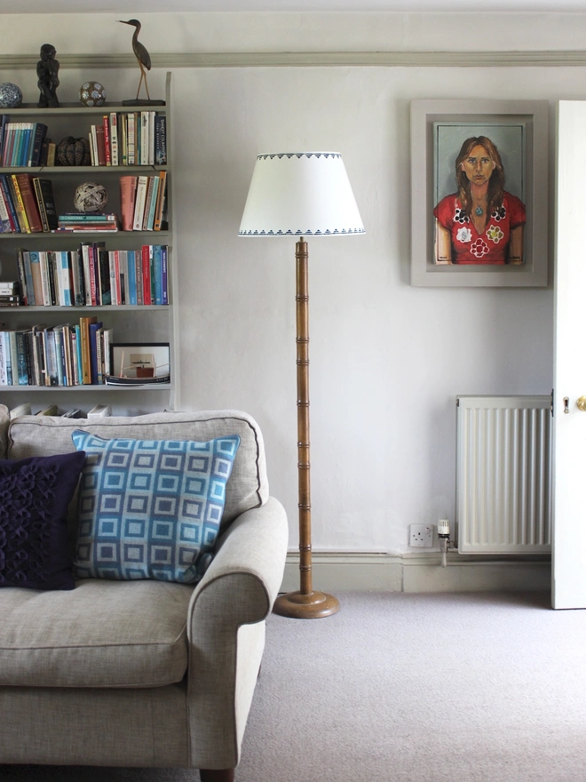 Living room interior full of sofa, bookshelf and portrait art, has a tall floor lamp fitted with a cream empire lampshade with a blue triangle pattern. This neutral lampshade with geometric detail adds a subtle colourful accent to the space.