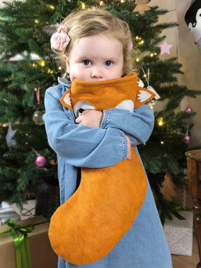A young girl wearing a denim dress stands in front of a Christmas tree hugging a handmade felt fox stocking.