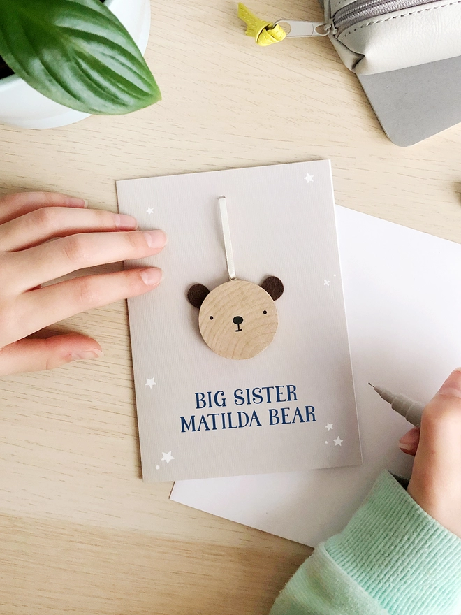 A grey greetings card with a small wooden bear keepsake and the words "Big sister Matilda bear" printed on is on a wooden desk. 