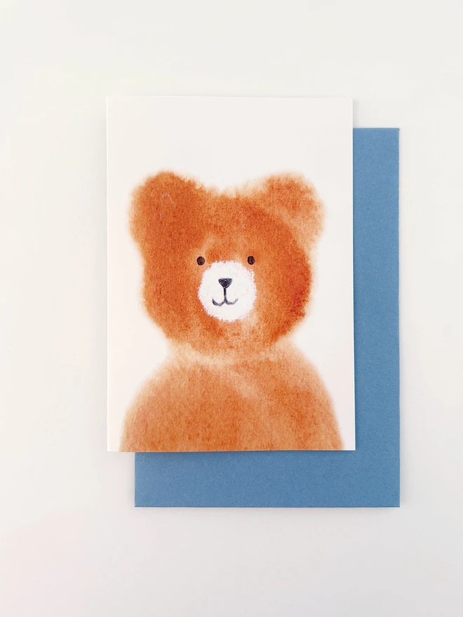 A greetings card featuring a hand painted brown bear cub. The cub is painted in watercolour with a white snout and a friendly smile. With a pale blue envelope