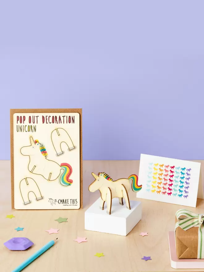 3D rainbow unicorn decoration and unicorn pattern greeting card and brown kraft envelope on a wooden desk in front of a lilac coloured background