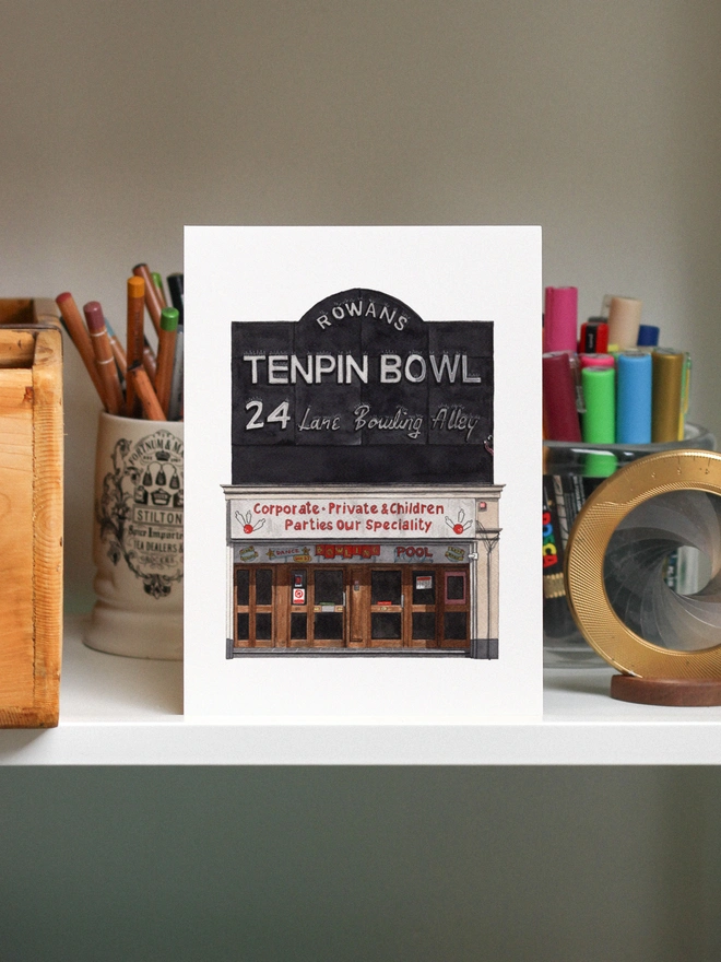 A watercolor print of the Rowans Tenpin Bowling  displayed on a shelf among various art supplies.