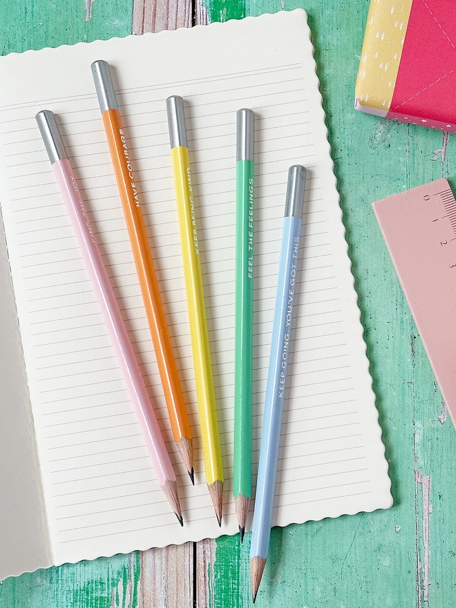 Five pastel coloured pencils, each with a silver end, lay on an open lined notebook on a green desk. 