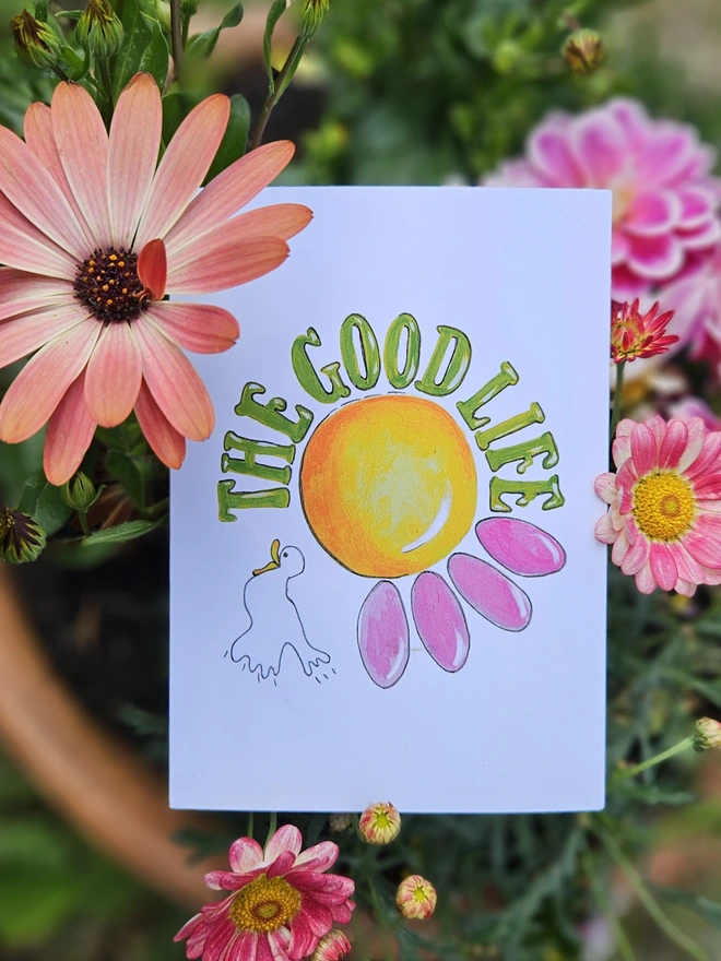 A white card with an illustration of a pink flower and a white bird with The Good Life written using a retro font in green. 