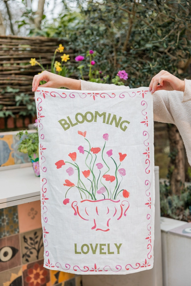 Linen teatowel with Blooming Lovely and floral pattern, held up against a garden background
