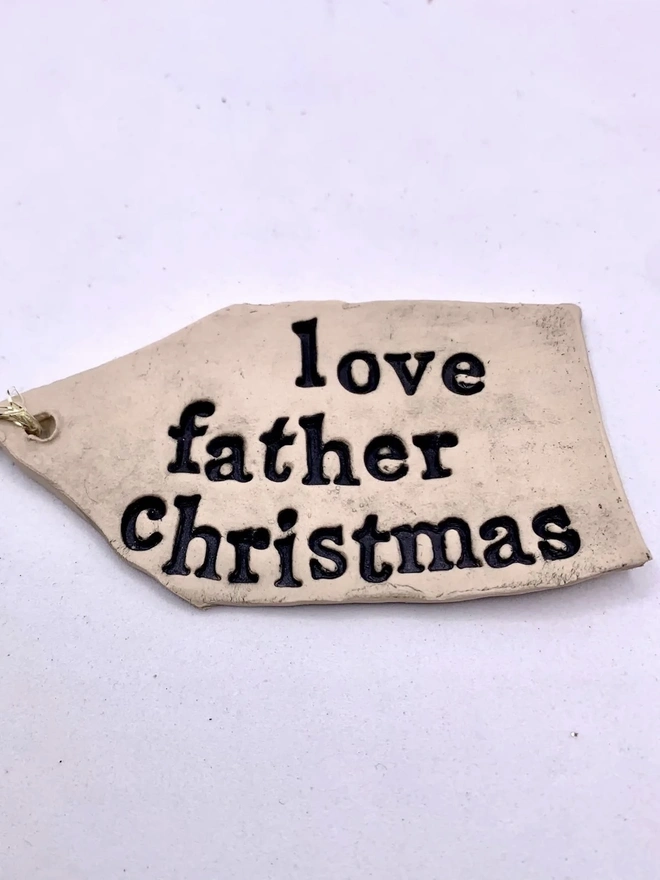 'Love Father Christmas' Handmade Ceramic Gift Tags, a ceramic gift tag attached sitting on a ale purple surface. 