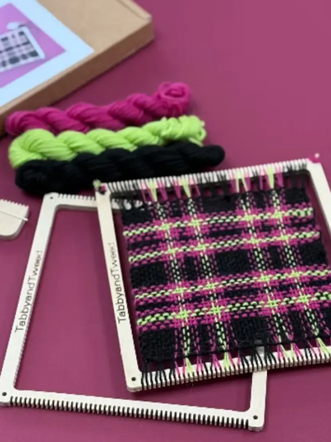 Square frame weaving loom kit with neon pink, green, and black tartan sample, yarn skeins, and wooden tools on a magenta background.
