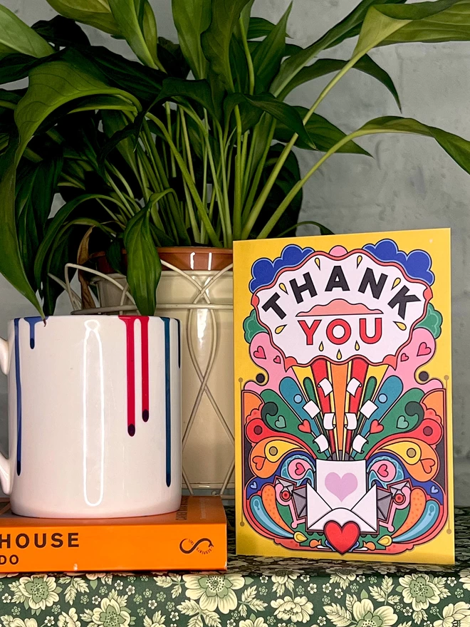 A vibrant yellow card with Thank You written boldly at the top, above a multi-coloured abstract design, sits on a shelf next to a pot plant, and a white mug with a paint drip design on top of an orange book. 