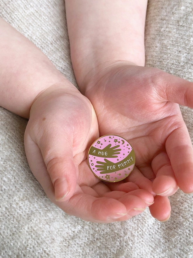 A child's hands are holding an enamel pin badge that has a hugging arms design and the words "A hug for Mummy".