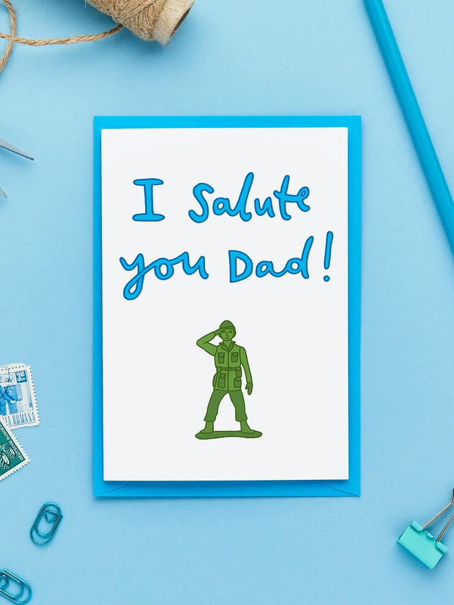 Greeting card featuring a green toy soldier illustration with the hand-lettered caption 'I Salute You Dad!', styled flat lay on blue background with stamps, string, pencil and paperclips.