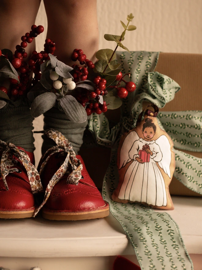 A Little inn asian dark skinned plush Christmas tree angel decoration placed next to a little girls red shoes