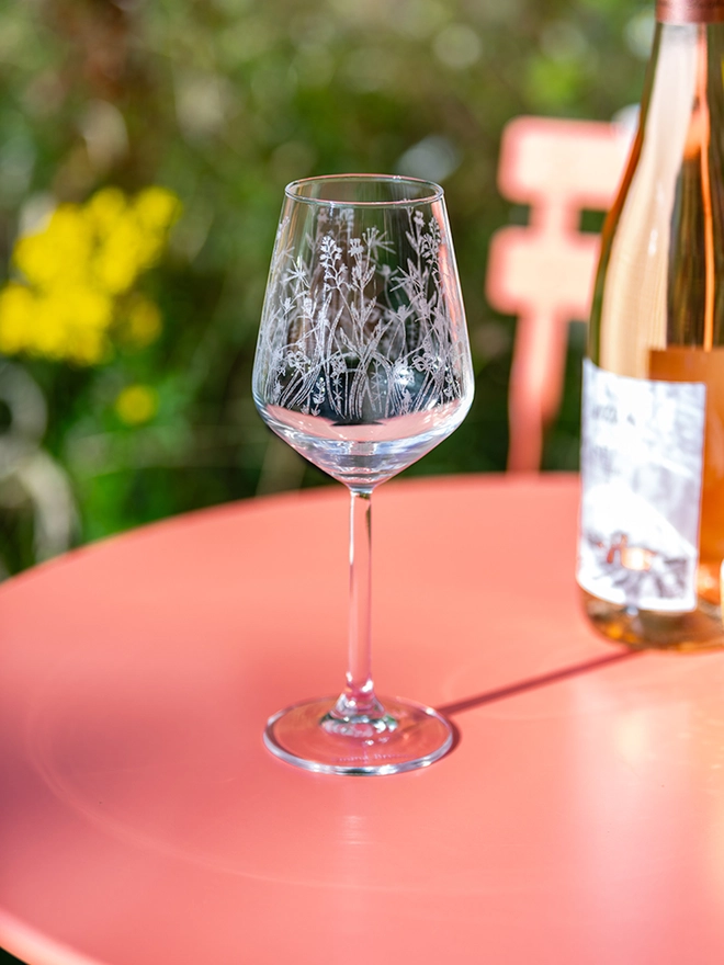 Meadow Wine Glass, an etched wine glass with a wildflower pattern sits on a bright pink patio table next to a bottle of rosé wine.