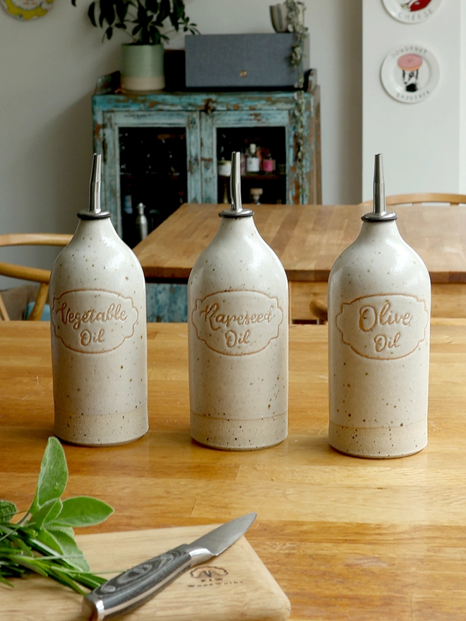 Three handmade stoneware oil pourers sat on a wooden table in a kitchen scene. The oil pourers feature the  words 'Vegetable Oil', 'Rapeseed Oil' and 'Olive Oil' and are glazed in an off-white glaze