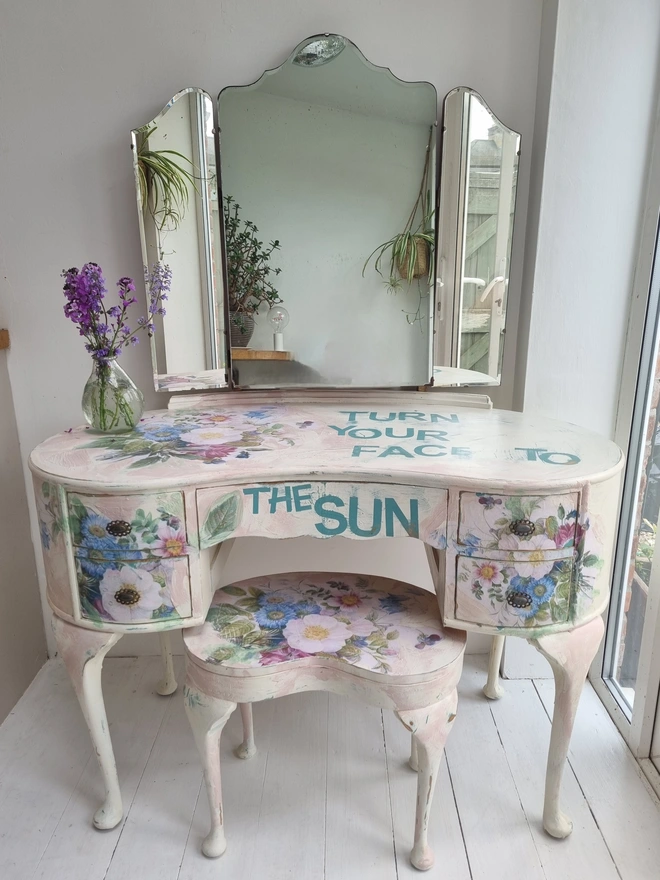 A painted dressing table in white and pale pink with large florals in blues and mauves and text in turquoise that reads 'turn your face to the sun'. The whole piece is distressed in the shabby chic style.
