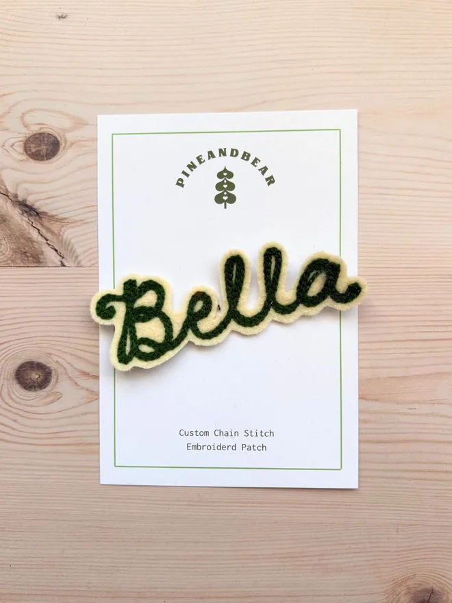 A chain stitched embroidered patch in cream and green spelling the word Bella, on a white presentation card