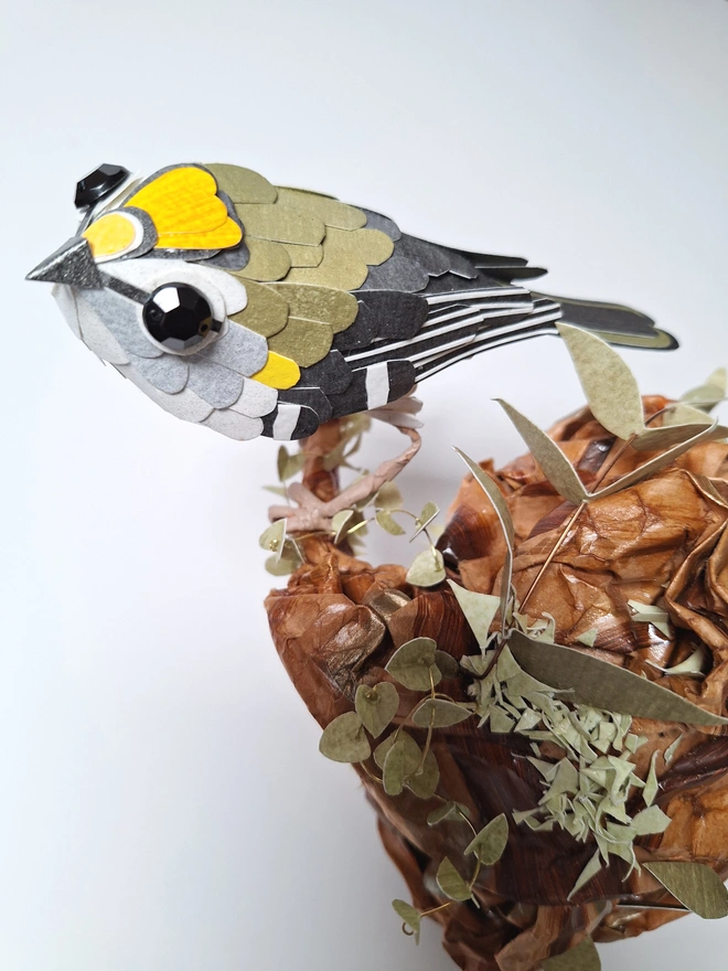 Firecrest paper bird sculpture, perched on a tree stump. Close up of the garden bird, made from handprinted papers.