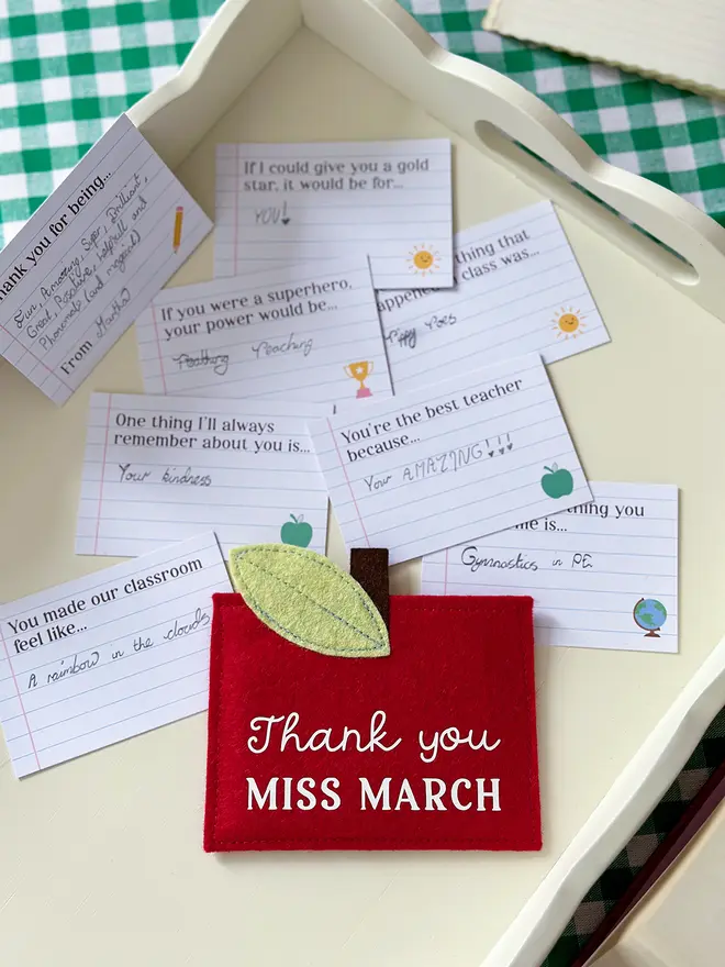 A handmade red felt apple-shaped envelope with "Thank you Miss March" written on the front. The heirloom apple envelope surrounded by completed prompt cards with handwritten messages from a child, displayed on a wooden tray. It's a unique personalised gift for teachers that is perfect for an end of term thank you.