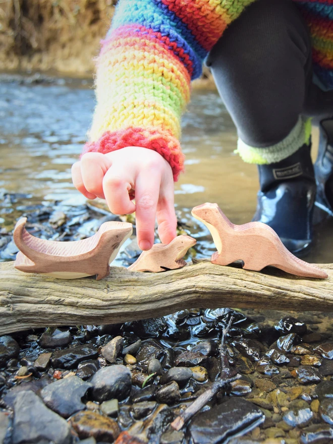 wooden otter toys being played with by the river by a child in a woolly jumper and wellies paddling in water