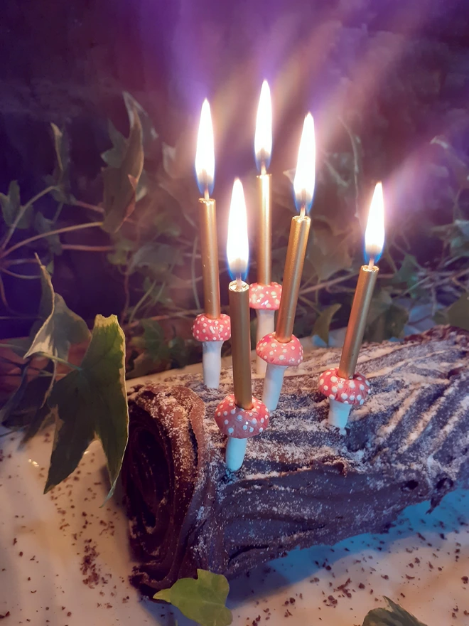 Angled close-up of five glowing gold cake candles in ceramic mushroom holders, decorating a chocolate yule log surrounded by ivy.