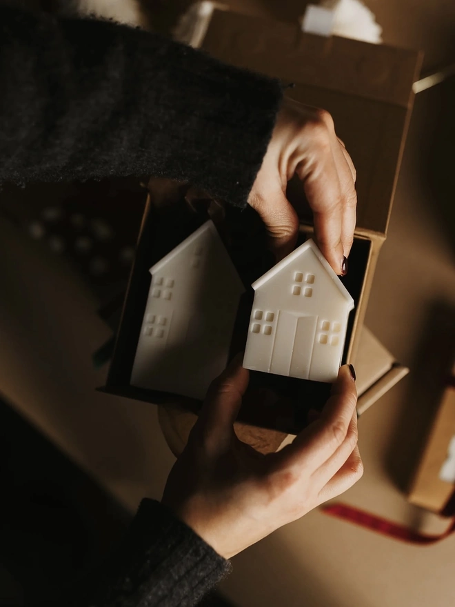 White Christmas Townhouse Candles (Set Of 3). Hands placing two cream house candles into a kraft gift box. The handmade candles arrive ready to give, making them a thoughtful housewarming or Christmas present.