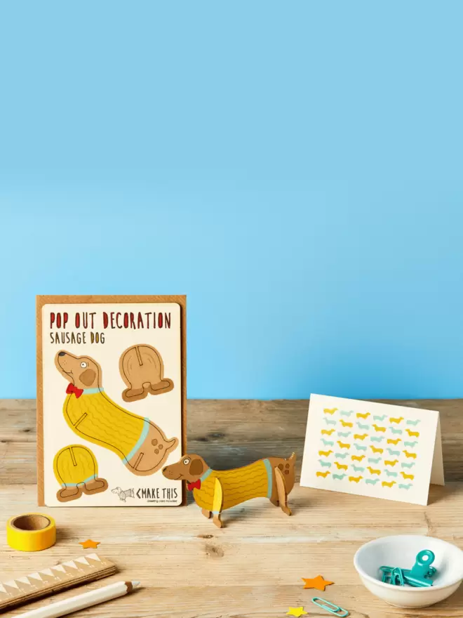 Sausage dog decoration and sausage dog greeting card with stationery items sitting on a wooden desk with blue background