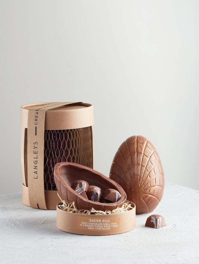 Extra Thick Milk Chocolate Easter Egg With Cornish Fudge, a chocolate egg sitting on a white surface. It is accompanied by another chocolate egg that is opened showing the chocolates within. 