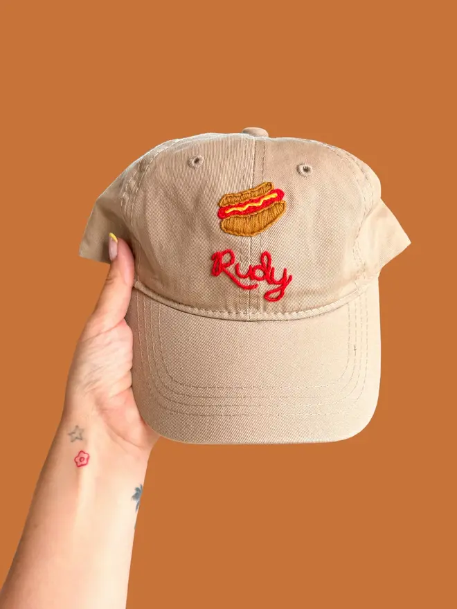 Hand Embroidered Kids Cap, a person holding a personalised cap against a colourful backdrop. It says a child's name. 