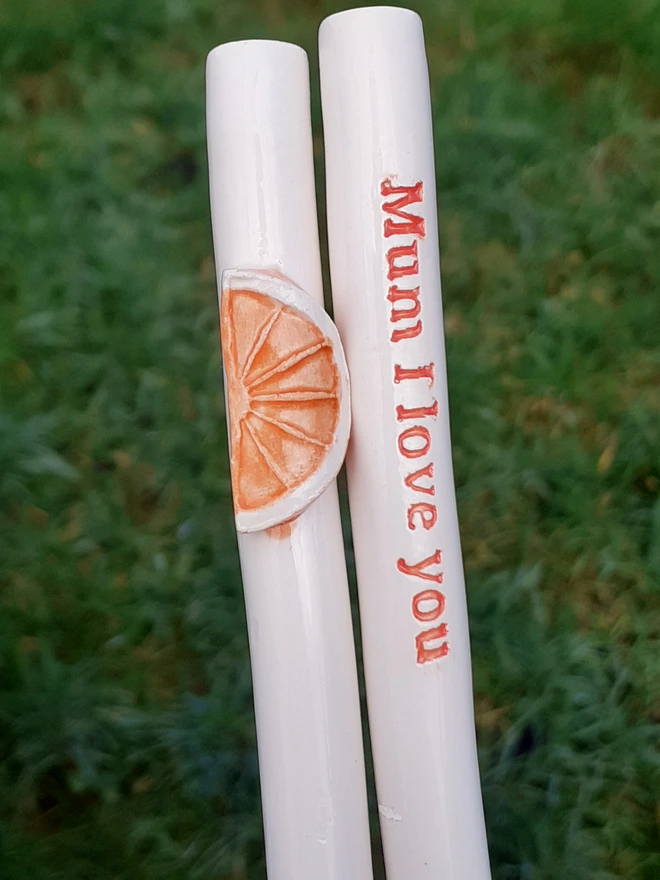 Two white ceramic drinking straws photographed outdoors on grass, one decorated with a raised orange slice motif and the other reading “Mum I love you” in red lettering.