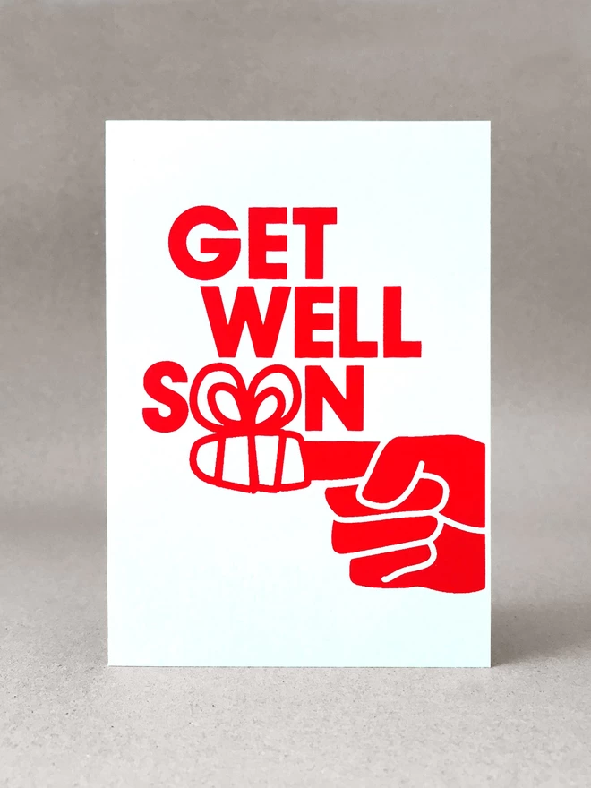 A finger wrapped in a bandage form the two o’s of the word SOON as part of this Get well soon card. Printed in red ink on a white portrait format card. Stood front on, in a light grey studio set.