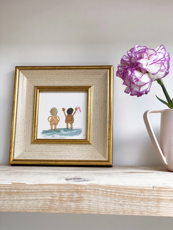 wooden shelf with purple and white carnation to the side and framed picture of two naked people cheerfully looking out to sea with pink goggles