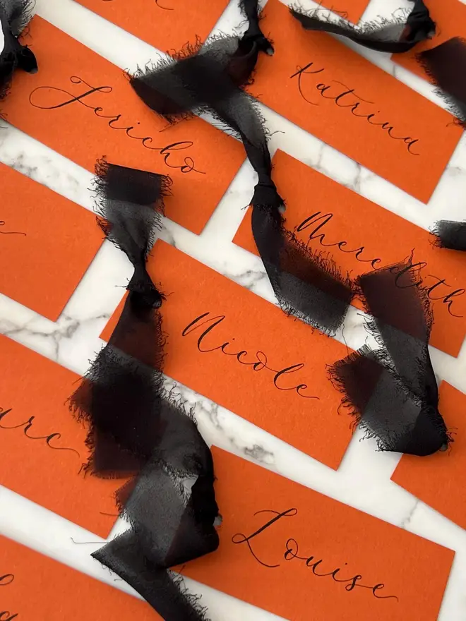 Personalised Calligraphy Rust Orange Place Card, orange place cards with black chiffon ribbons grouped together on a white surface. 
