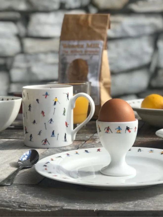 Ski Chain Breakfast Set. A side plate, a bowl, an egg cup, an espresso cup, and a saucer. All are a plain white with a pattern of small, multicoloured skiers. They are set against a stone background on a table.
