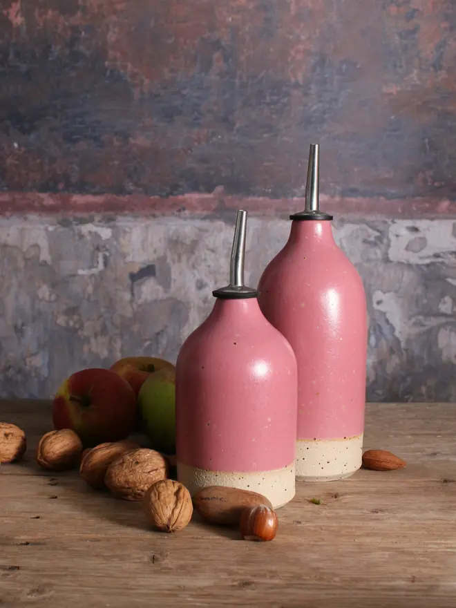 Small and large ceramic oil pourers in fuchsia sat on a wooden surface amongst scattered seasonal fruits. 
